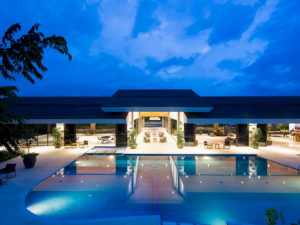 A luxurious, modern building with a large, illuminated swimming pool in front, surrounded by outdoor seating, under a deep blue evening sky.