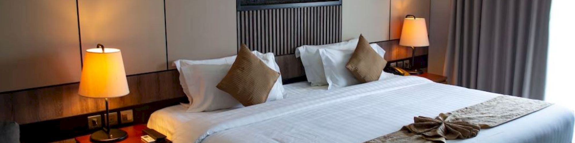 A hotel room features a large bed with white linens, brown accents, bedside tables with lamps, neutral tones, and a decorative wall art piece.