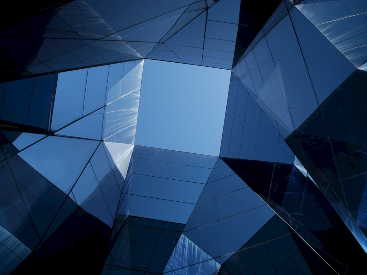 The image shows a view looking up through a geometric structure of reflective glass panels with a blue sky visible in the center.
