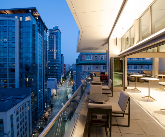 A modern balcony with seating overlooks a city street with high-rise buildings at dusk.