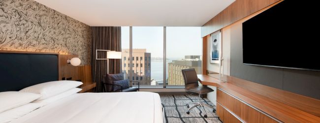 A modern hotel room with a large bed, wall-mounted TV, desk, and a window view of buildings, showcasing a sleek, minimalist design.