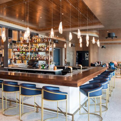 The image shows a modern bar with blue stools, a wooden counter, shelves stocked with bottles, and hanging pendant lights in a stylish space.