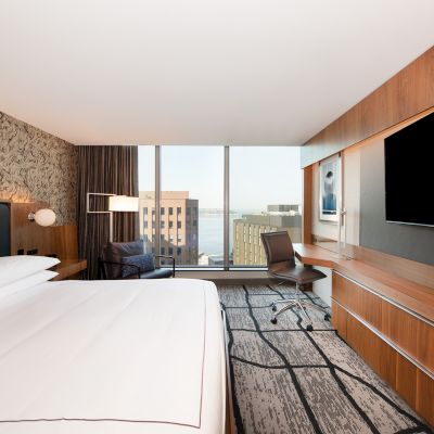 A modern hotel room with a large bed, TV, desk, and window view of buildings outside, featuring stylish decor and lighting.