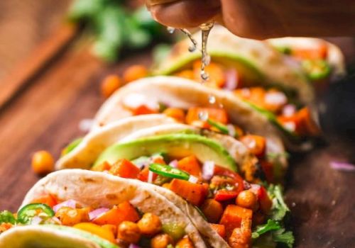 Tacos filled with chickpeas, avocado, and veggies are arranged on a board. A hand squeezes lime juice over them, with more limes nearby.