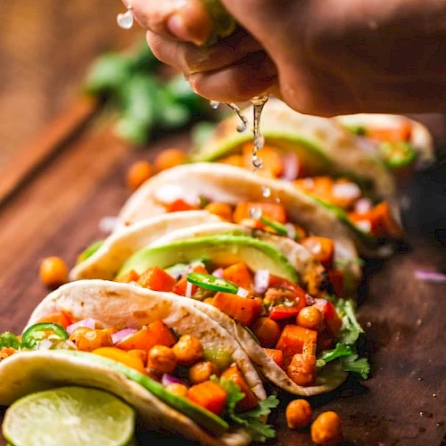 Tacos filled with chickpeas, avocado, and veggies are arranged on a board. A hand squeezes lime juice over them, with more limes nearby.