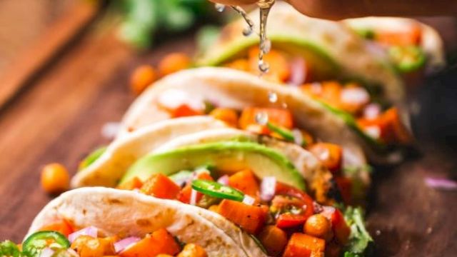 Tacos filled with chickpeas, avocado, and veggies are arranged on a board. A hand squeezes lime juice over them, with more limes nearby.