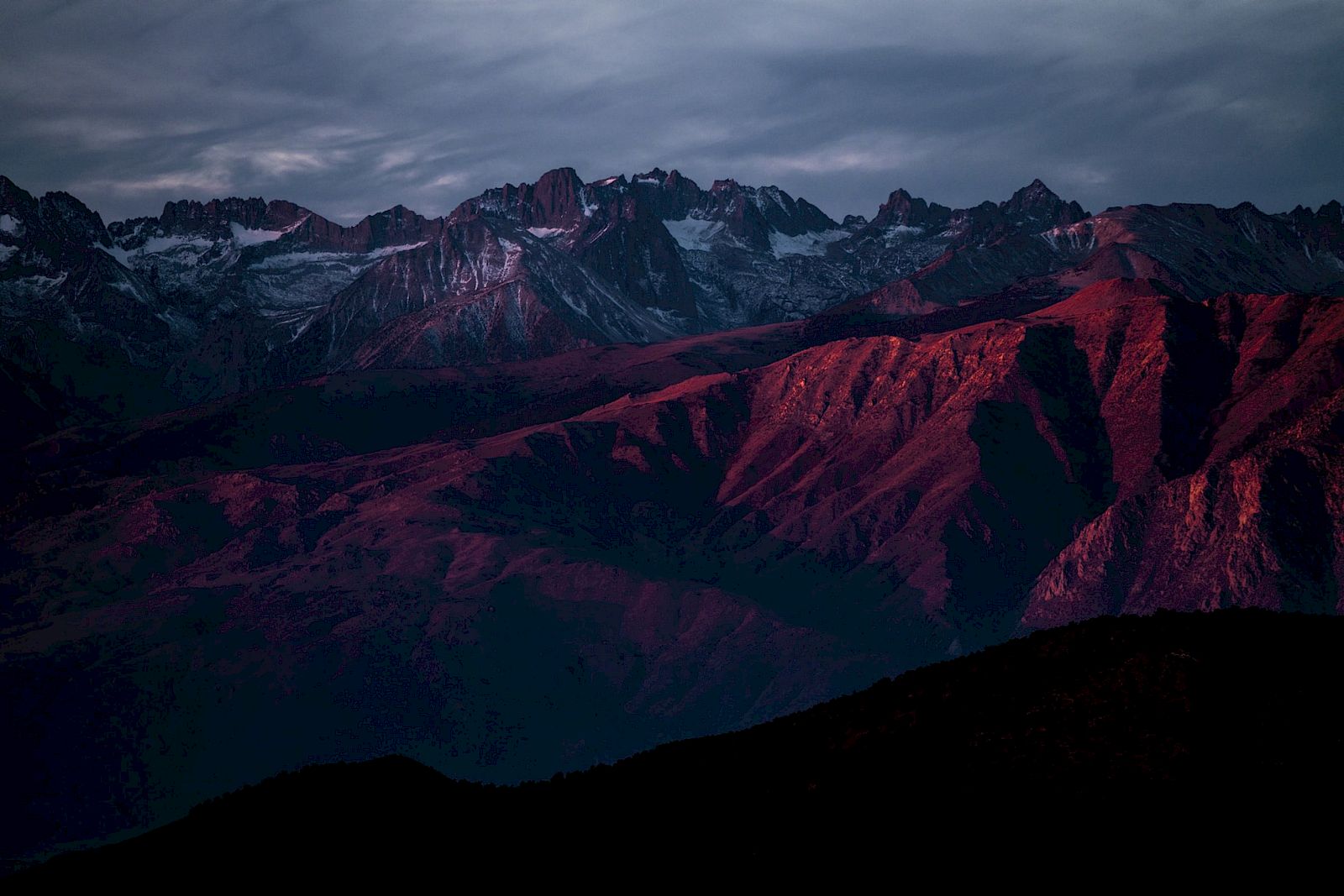 A mountain range under a dramatic sky, with the peaks bathed in a reddish hue, creating a stunning contrast against the dark surroundings.