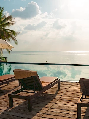 A serene beach scene with lounge chairs, umbrellas, palm trees, and an infinity pool overlooking the calm ocean under a partly cloudy sky.