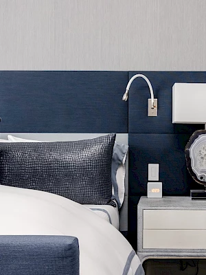 A modern bedroom features a bed with blue and gray bedding, a nightstand with a lamp and decor, and a light gray wall.
