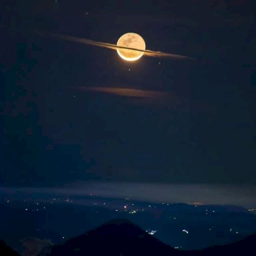 A full moon appears with a thin cloud resembling Saturn's rings, above a dark landscape with distant city lights.