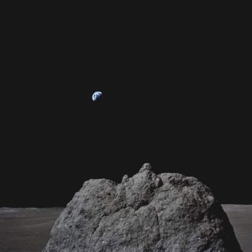 A barren moon surface with a large rock in the foreground and Earth visible in the distant, dark sky.