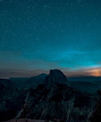 A night sky filled with stars over a mountain range, with a silhouette of a prominent peak and a glow on the horizon.