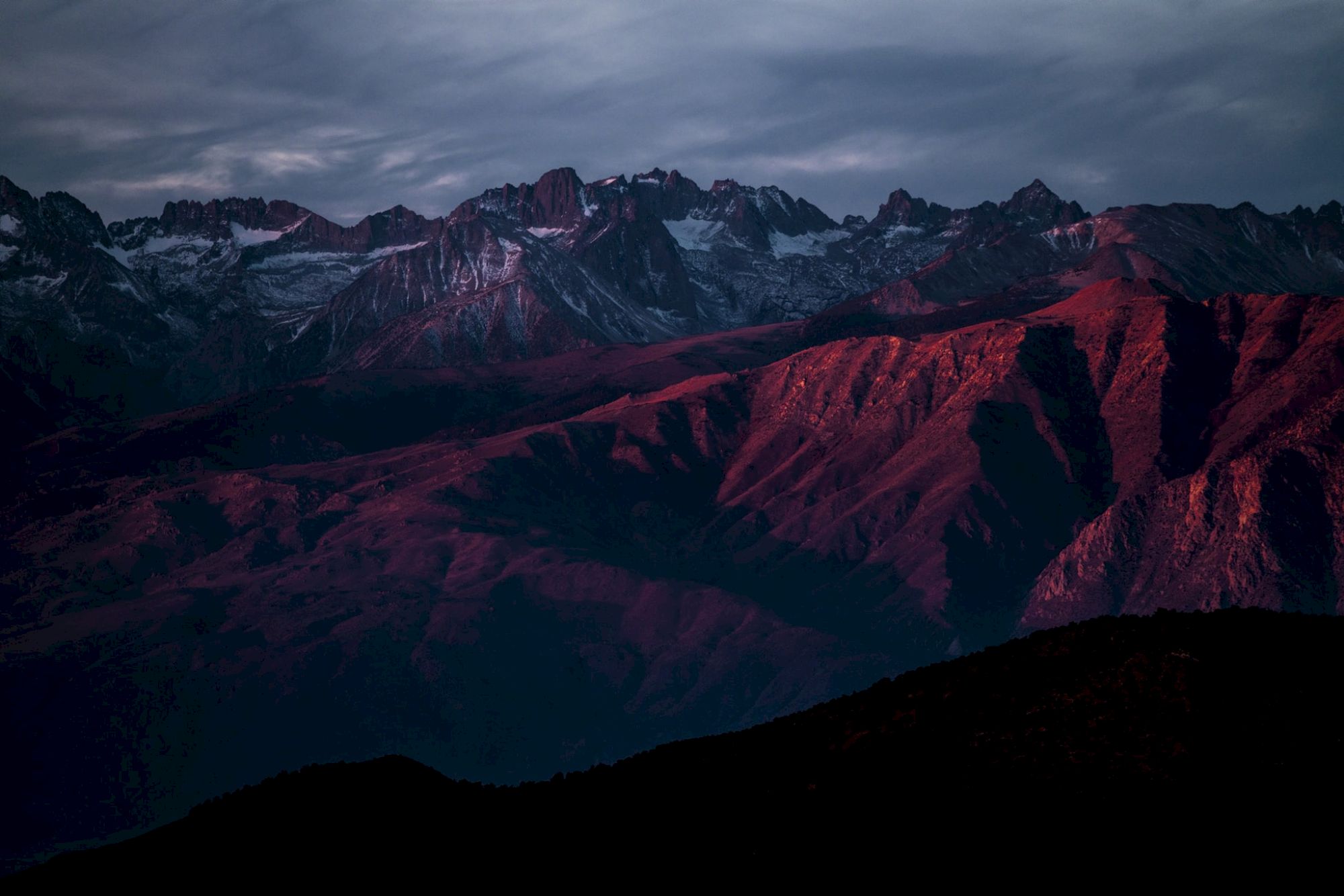 A mountain range at sunset, with peaks highlighted in deep red hues against a moody sky.