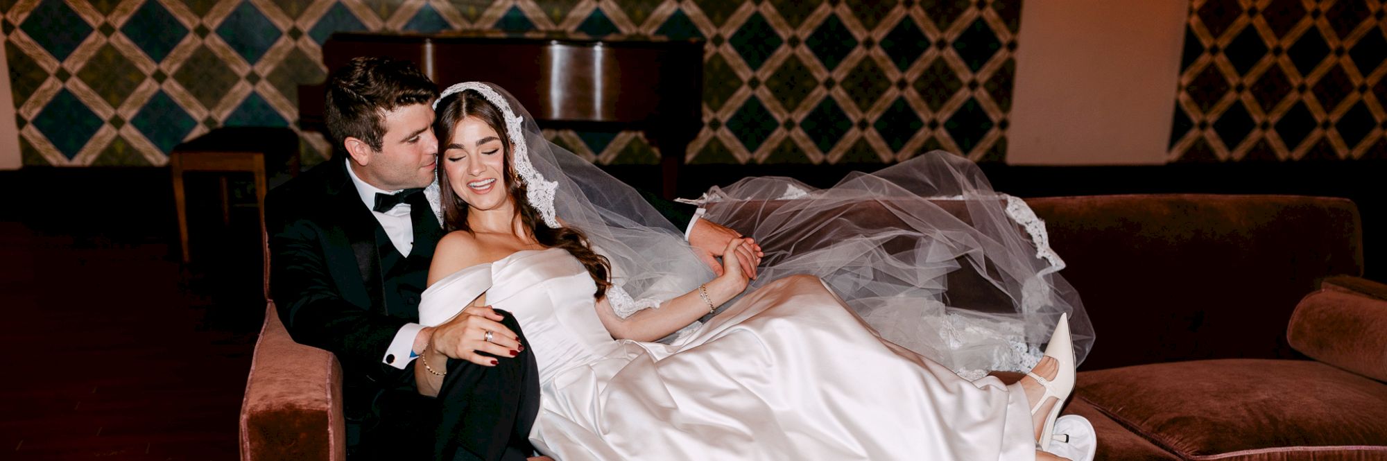 A bride and groom are seated on a couch. The bride wears a white dress and veil, while the groom is in a tuxedo, posing affectionately.