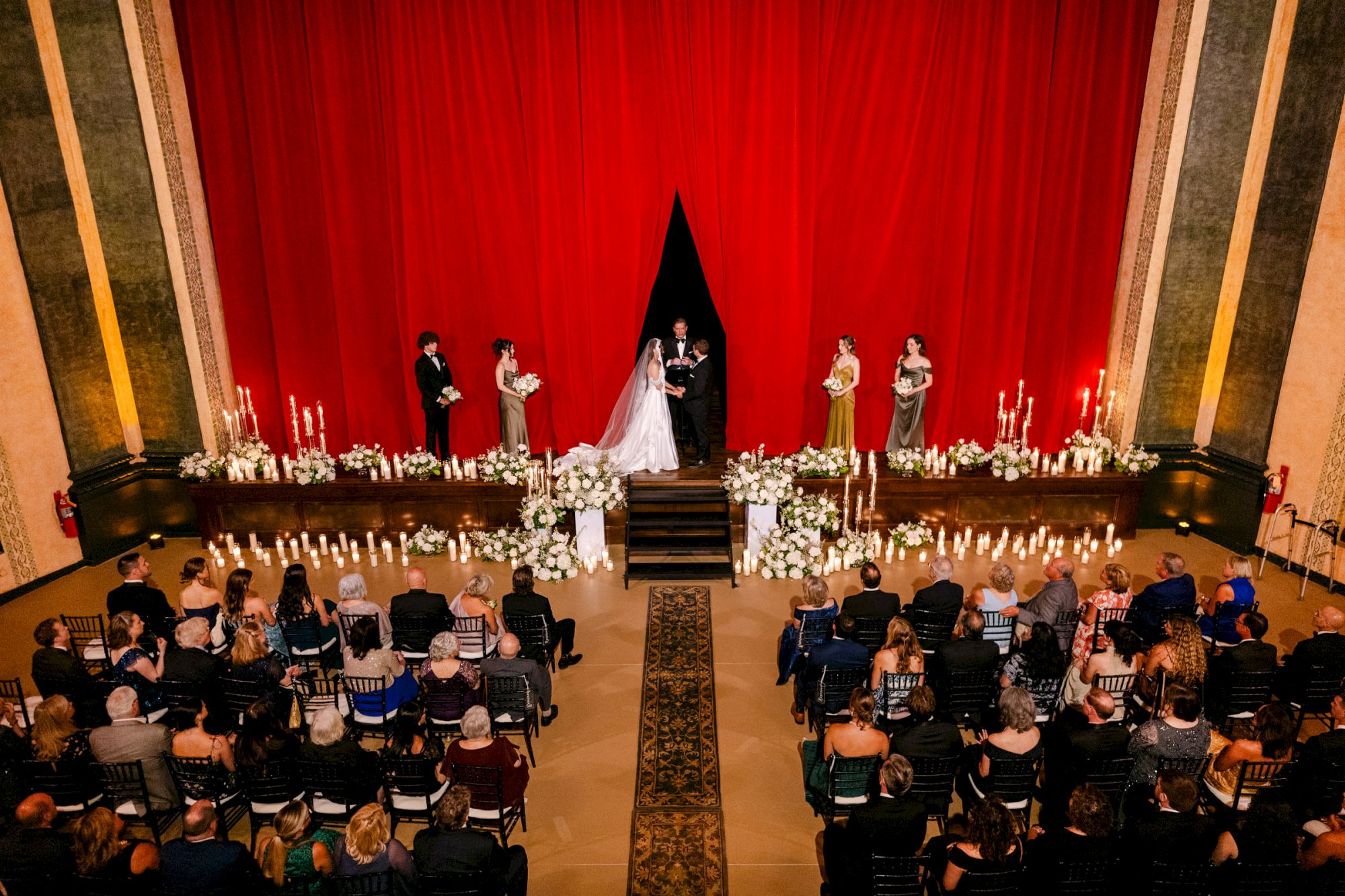 A wedding ceremony is taking place on a stage with a red curtain backdrop, surrounded by flowers and candles, attended by many guests.