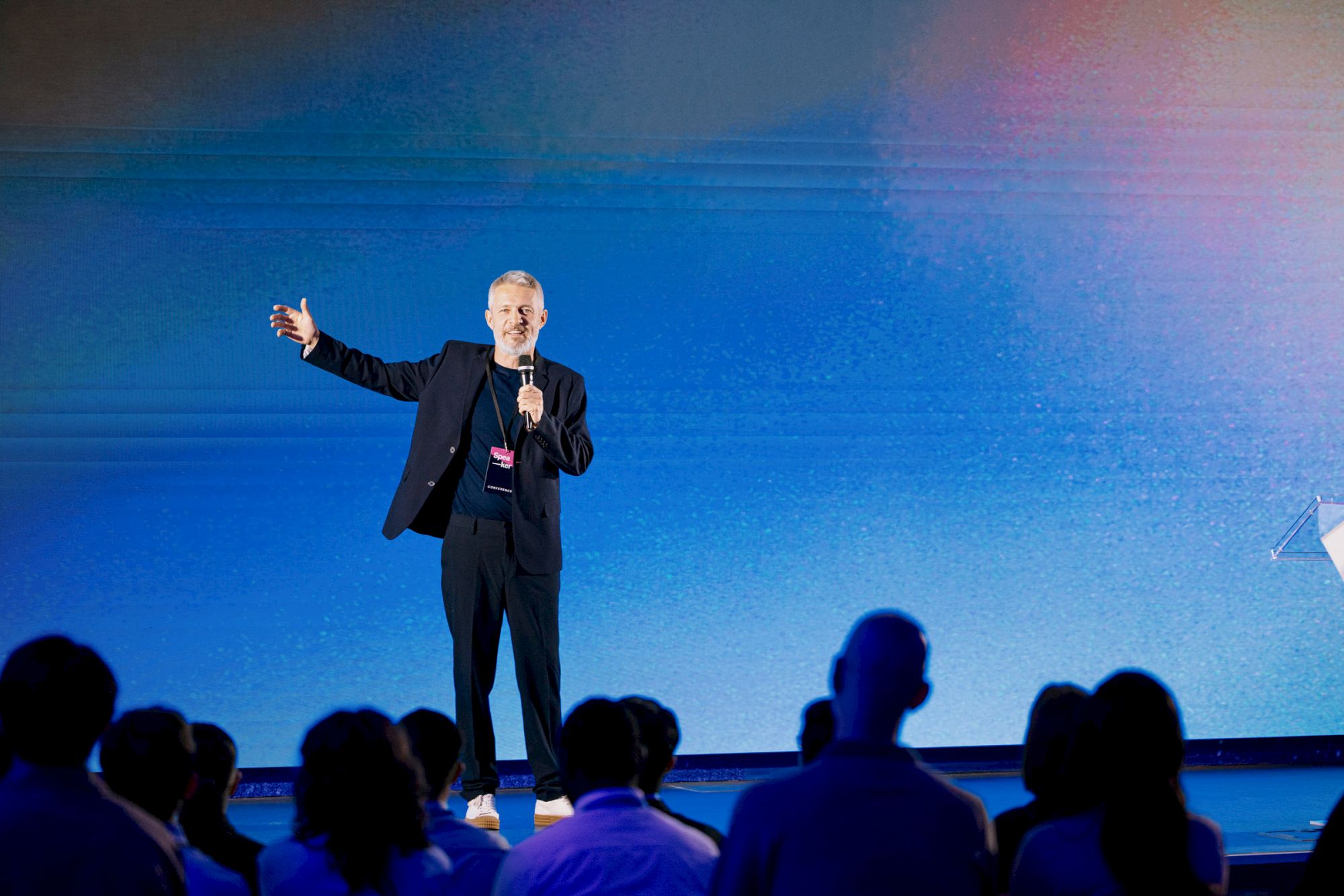A person in a suit is speaking on stage with a microphone, gesturing toward an audience seated in front of a blue background.