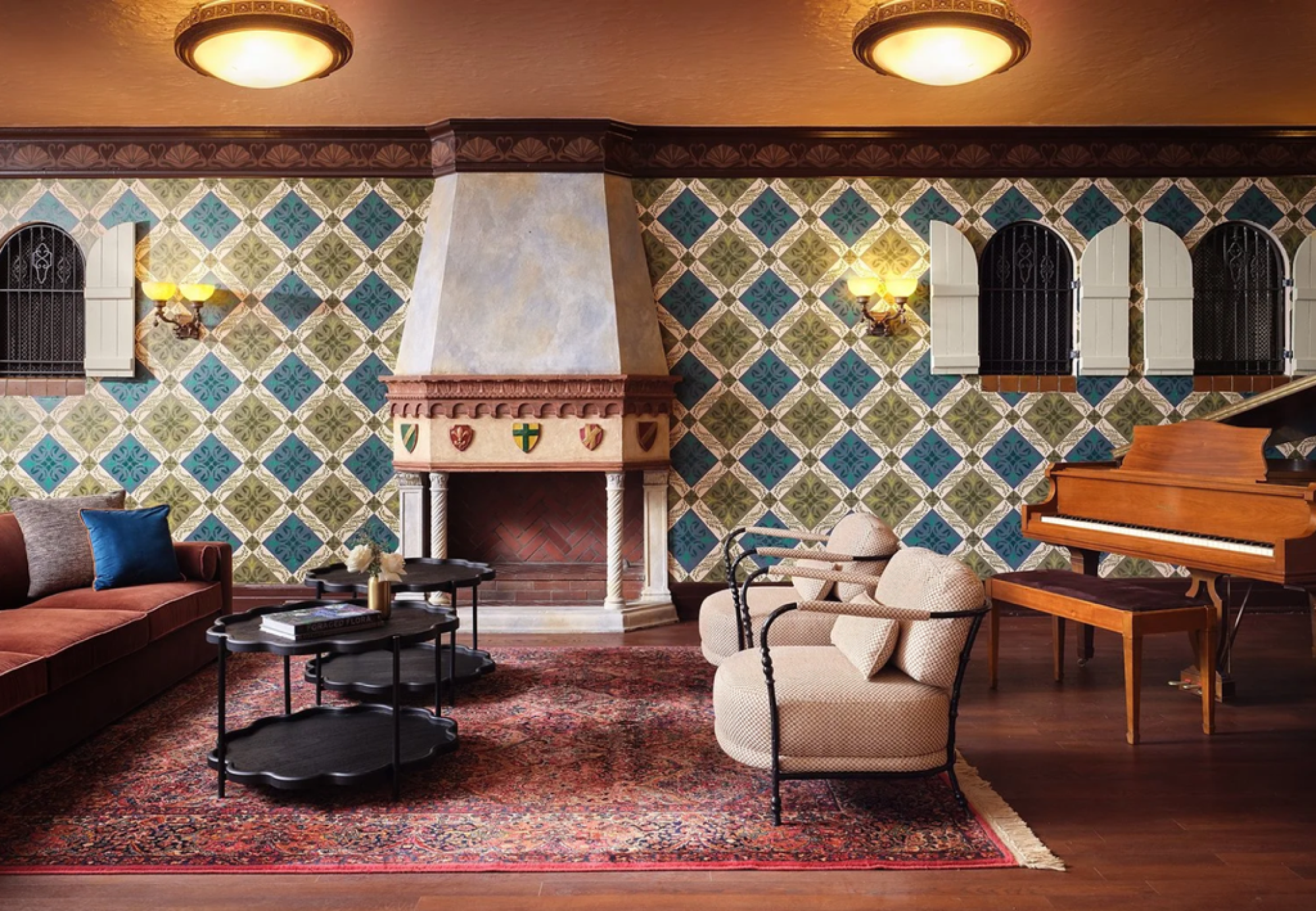 A cozy living room with a fireplace, patterned wallpaper, seating area, coffee tables, and a piano on a wooden floor with a decorative rug.