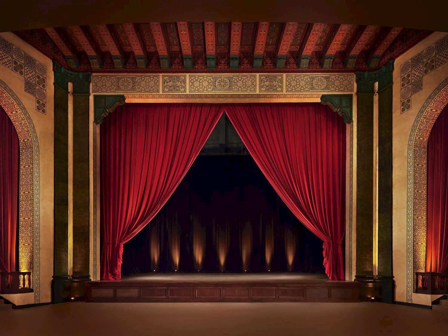 A grand theater stage with red curtains, ornate gold and green decor, and soft backlighting in a historic, elegant interior.