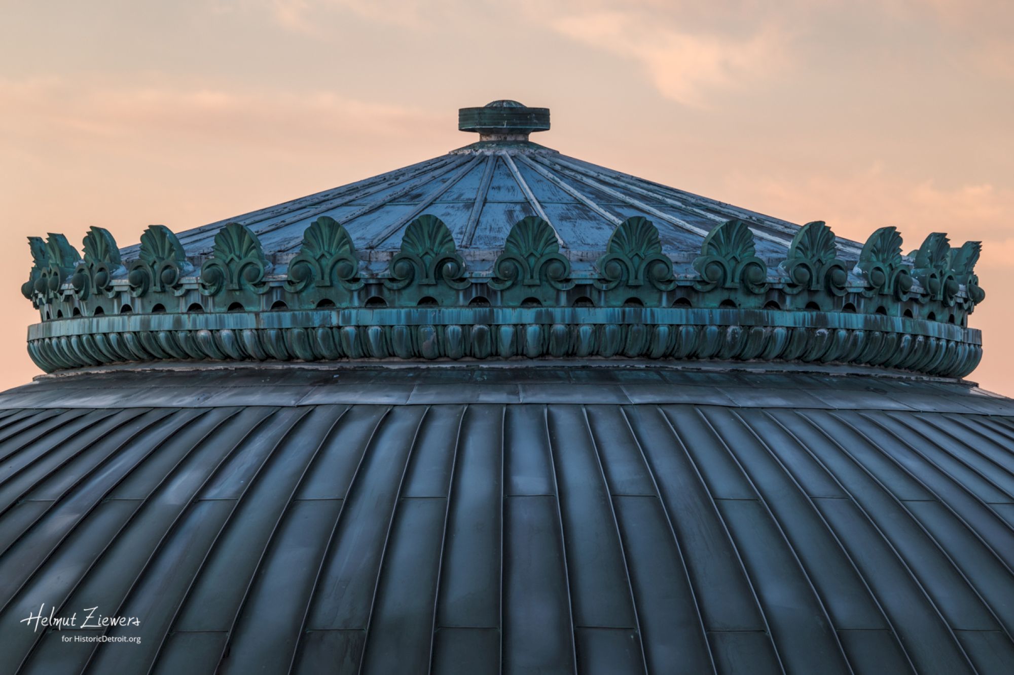 The image shows the ornate top of a domed structure, with intricate detailing and patina, under a soft, pastel sky.