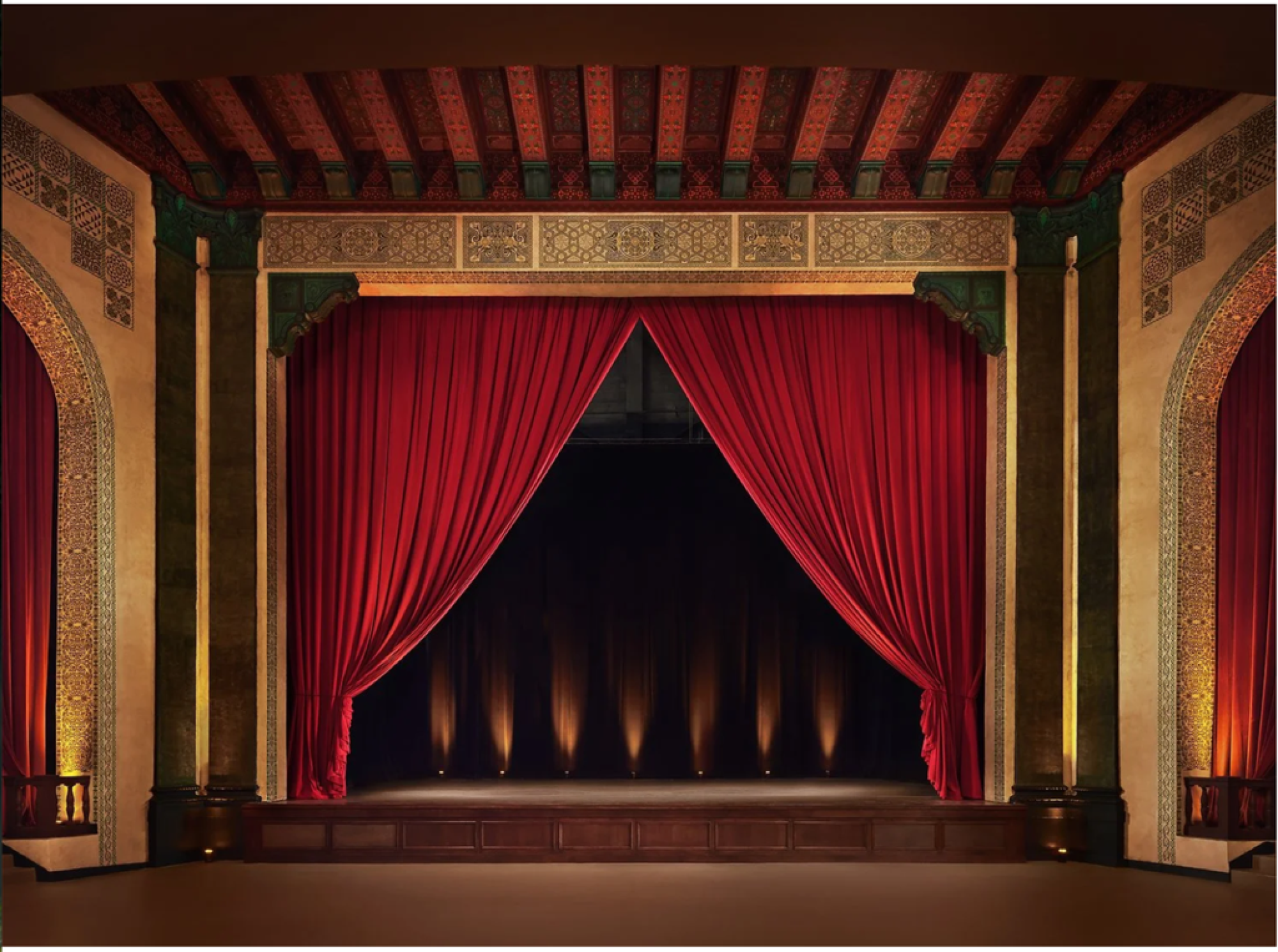 An ornate theater stage with red curtains drawn to the sides, revealing a dark backdrop with subtle lighting below the stage.