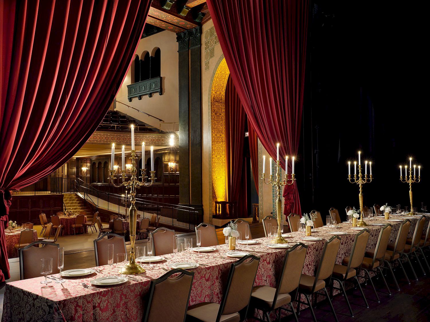 A lavish banquet hall with red velvet curtains, chandeliers, and a set dining table with candles and glasses, lit for an event.