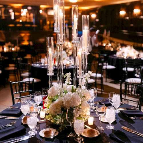 A beautifully set dinner table with elegant glassware, floral centerpiece, and candles in a dimly lit banquet room.