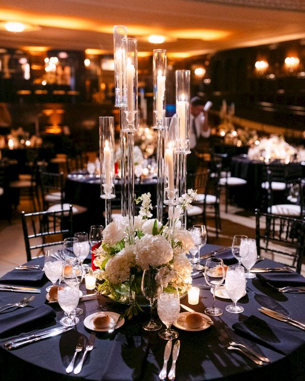 A beautifully set dinner table with elegant glassware, floral centerpiece, and candles in a dimly lit banquet room.