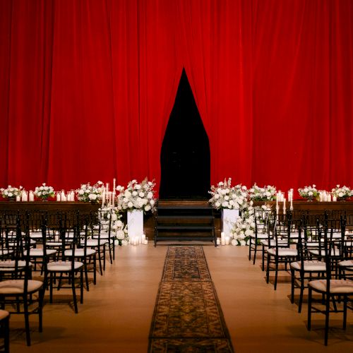A wedding ceremony setup with rows of chairs, decorative candles, and flowers, in front of a red curtain on a carpeted aisle.