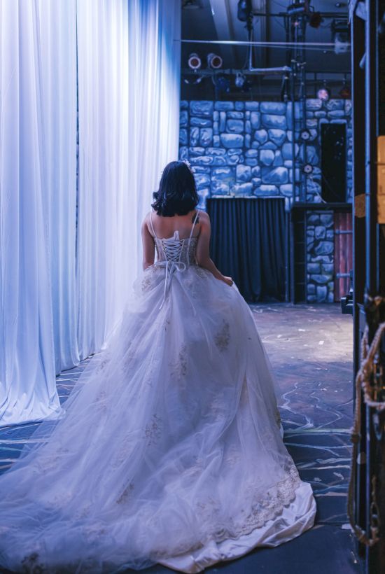 A person in a wedding dress stands backstage, with draped curtains and stage equipment around, creating a dramatic scene.