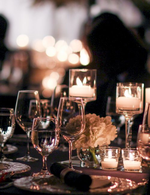 The image shows a dimly lit elegant dinner table with wine glasses, candles, and floral decor, creating a sophisticated atmosphere.