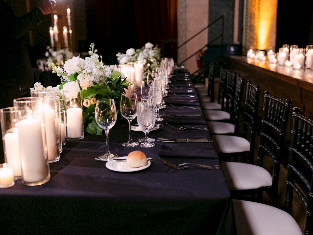 A dimly lit dinner table with black chairs, set for an event, featuring candles, flowers, glassware, and a single bread roll per setting.