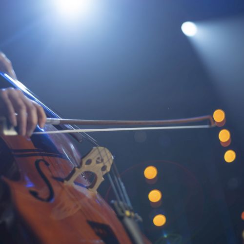 A musician is playing a cello under stage lights, with a blurred background of circular light spots.