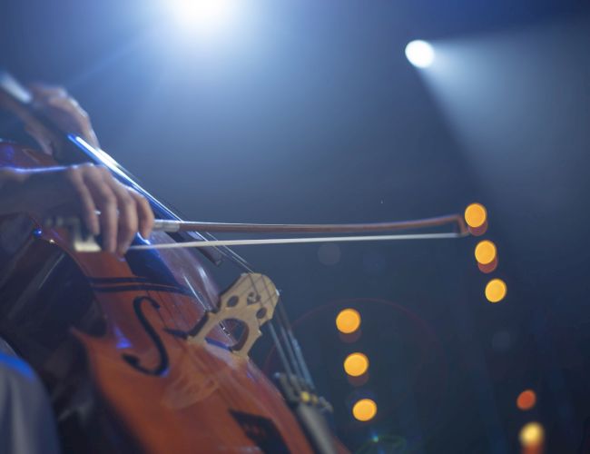 A musician is playing a cello under stage lights, with a blurred background of circular light spots.