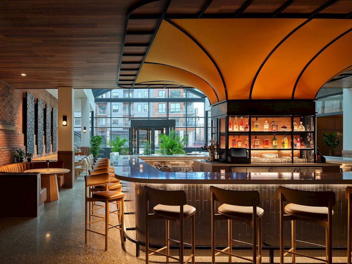 This image shows a modern bar with high chairs, an orange canopy, shelves with bottles, and seating areas with tables and benches.