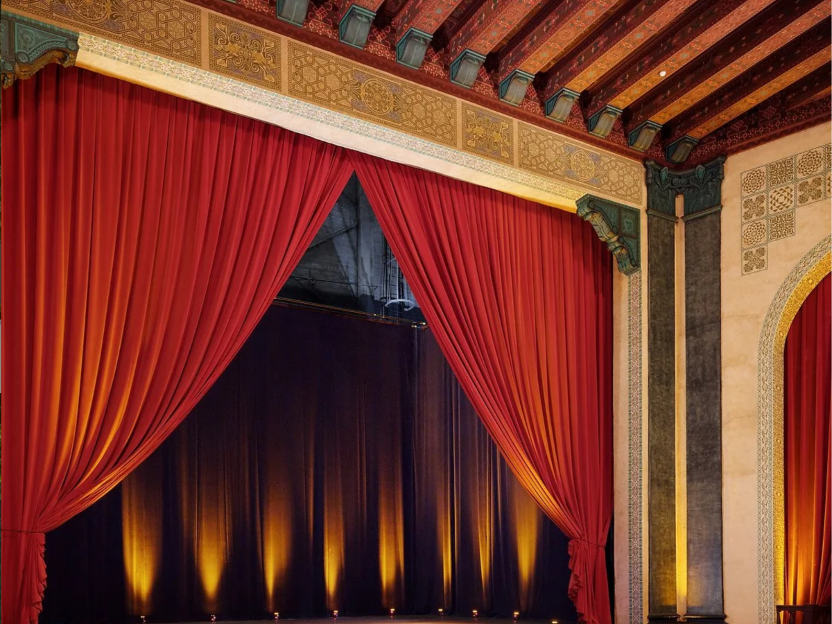 The image shows a theater stage with red curtains and ornate decorations, illuminated by warm lighting from below on the stage.