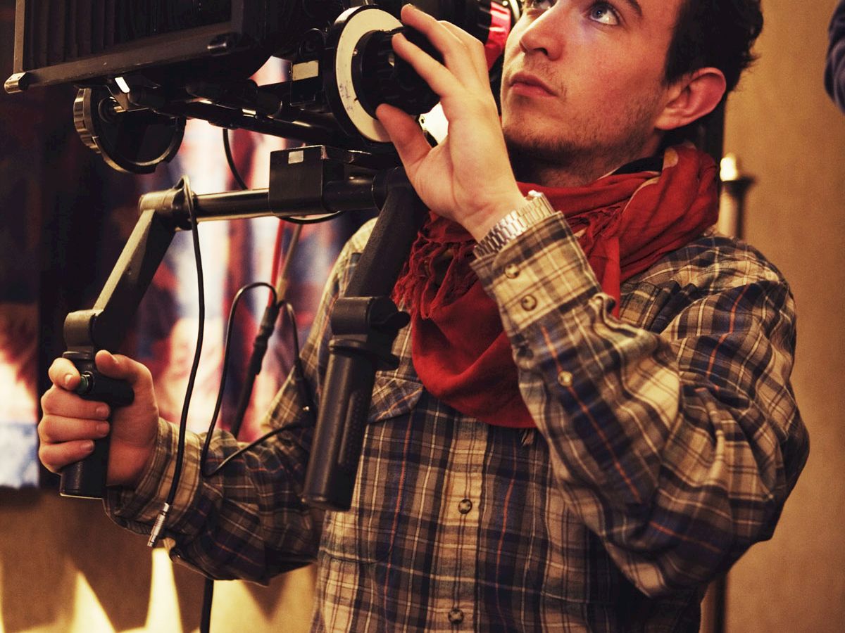 A person operating a professional camera, wearing a plaid shirt and red scarf, focused on filming in an indoor setting.