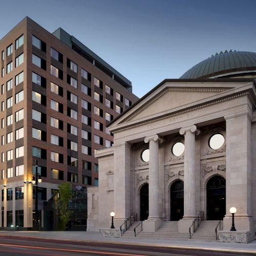 A modern brick office building beside a historic stone courthouse with a domed roof, steps, and circular windows, at dusk.