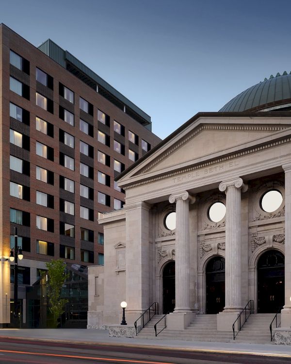 A modern brick office building beside a historic stone courthouse with a domed roof, steps, and circular windows, at dusk.