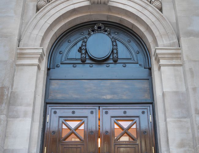 A tall arched doorway with ornate stonework, wooden double doors, brass handles, and decorative metal accents above and around the entrance.