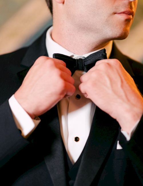 A person adjusting a bow tie while wearing a classic black tuxedo, with a blurred background.