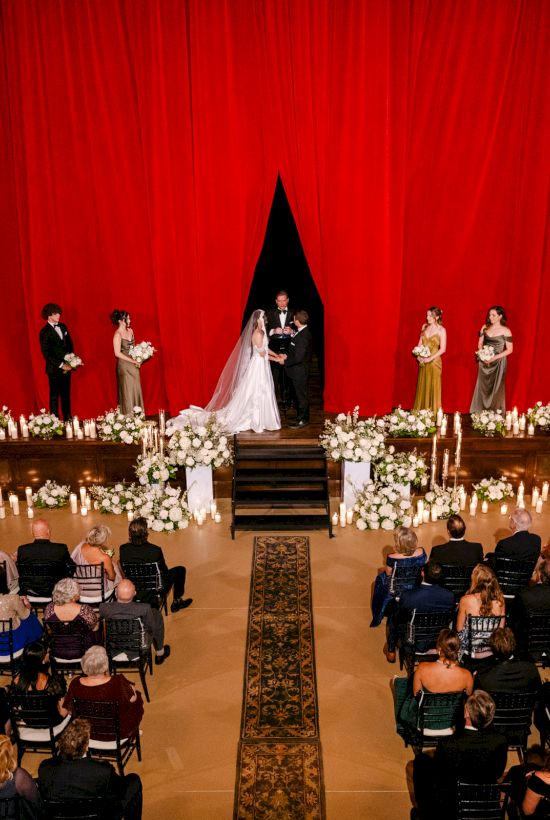 A wedding ceremony on a grand stage with a red curtain, floral decorations, and guests seated in rows.