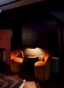 A cozy corner with two orange chairs, a small round table with a lamp, near a brick fireplace and a patterned rug on the wooden floor.