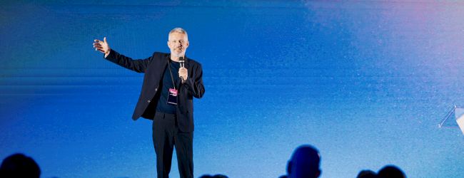 A person is speaking on stage with a microphone, gesturing to an audience against a blue background.