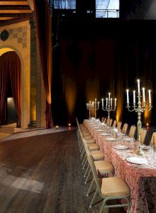 A long, elegant banquet table with a lace tablecloth and candles in a grand, dimly lit hall with tall arches and rich drapes.