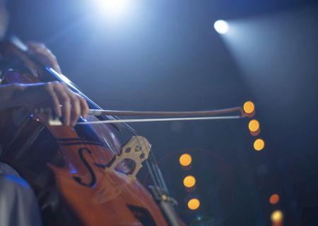 A close-up of a person playing a cello under stage lights, with a bokeh effect in the background.