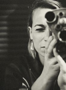 A person is looking through an old-fashioned film camera, focusing intensely. The background is dark and blurred for dramatic effect.