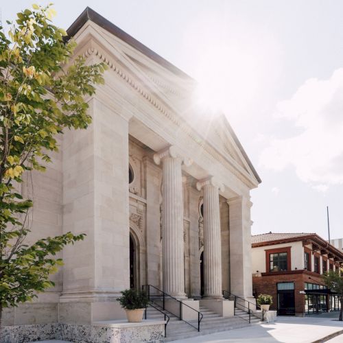 A sunlit neoclassical building with tall columns, a tree on the side, and brick pavement in the foreground.