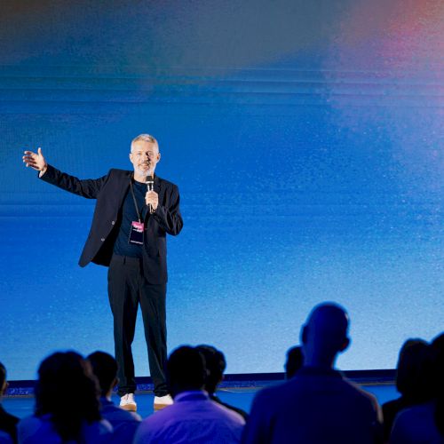A person is speaking on a stage with a microphone in front of an audience, set against a blue gradient background.