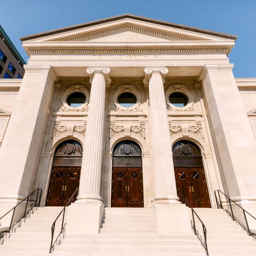 The image shows a grand building with columns, three arched doorways, and steps leading up, suggesting a neoclassical architectural style.