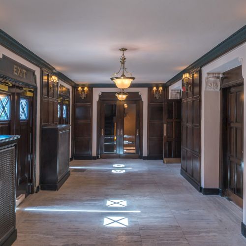 A hallway with wood paneling, chandeliers, and wall sconces, featuring elevator doors and an exit sign on the left.